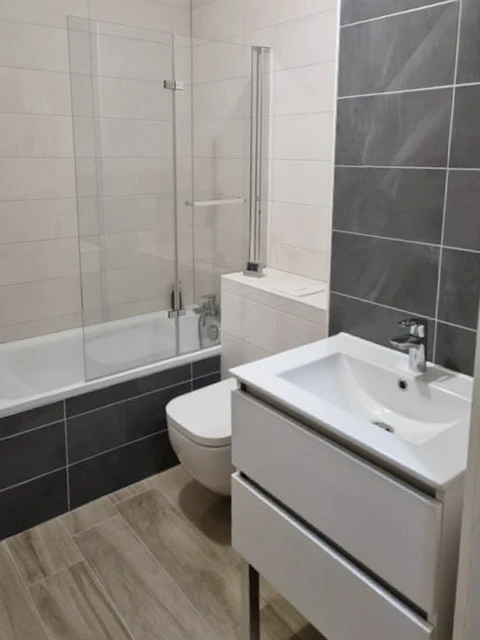 Salle de bain moderne compacte avec baignoire d'angle équipée d'un pare-douche en verre, WC suspendu blanc, meuble vasque suspendu blanc avec tiroirs et robinetterie chromée