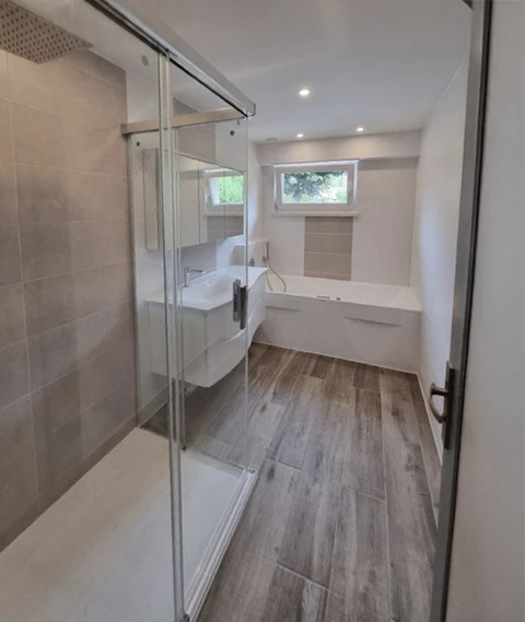 Salle de bain moderne entièrement rénovée avec douche à l'italienne, cabine de douche en verre trempé, vasque suspendue blanche, baignoire encastrée près d'une fenêtre